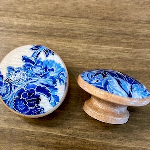 Könnte beinhalten: Zwei dekorative Schrankknöpfe. Einer ist rund, der andere oval, beide mit hellem Holzsockel. Die Oberseiten weisen ein blaues Blumenmuster auf weißem Hintergrund auf. Die Knöpfe sind für Möbel und Heimdekoration bestimmt.