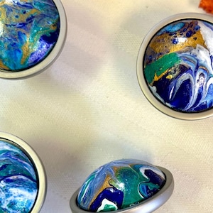 Könnte beinhalten: Vier Schrankknöpfe mit einem einzigartigen, abstrakten Design. Jeder Knopf hat eine runde, silberfarbene Basis und ein farbenfrohes, wirbelndes Muster in Blau-, Grün-, Gold- und Weißtönen. Ein Knopf ist aufrecht positioniert und zeigt seinen pilzförmigen Stiel.
