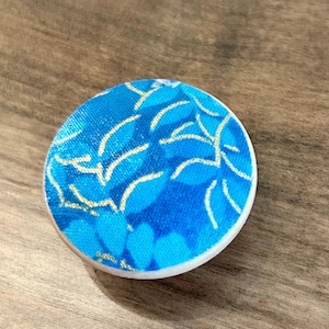 May include: A round, blue and white decorative button with a floral pattern. The button has a white edge and a blue fabric top with a gold leaf design. The button is on a wooden surface.