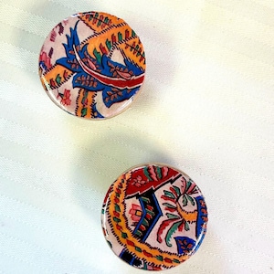 May include: Two round, decorative, wooden buttons with a paisley pattern. The buttons are covered in a fabric with a colorful paisley design featuring shades of blue, red, yellow, and green.
