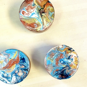 Könnte beinhalten: Drei runde Untersetzer aus Kunstharz mit Marmoreffekt. Die Untersetzer zeigen Wirbel aus blauen, weißen, goldenen und kupferfarbenen Farbtönen. Eine einzigartige und künstlerische Ergänzung für jede Inneneinrichtung.