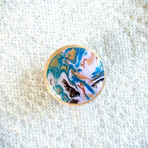 May include: A round wooden coaster with a colorful abstract design. The design features swirls of blue, gold, white, and brown, with a glossy finish. The coaster is approximately 3 inches in diameter.