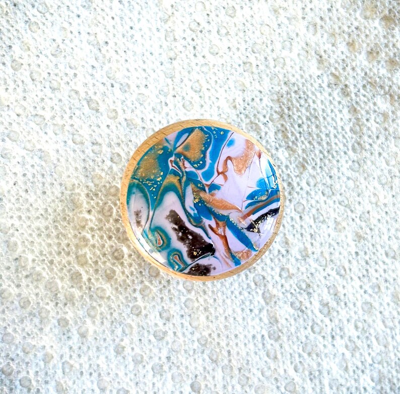 May include: A round wooden coaster with a colorful abstract design. The design features swirls of blue, gold, white, and brown, with a glossy finish. The coaster is approximately 3 inches in diameter.