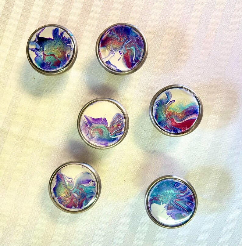 May include: Six round, silver-toned drawer knobs with a white background and abstract purple, blue, and green swirls.