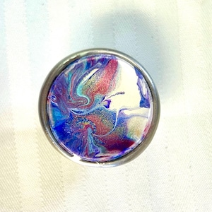 Puede incluir: Un pomo redondo de color plateado con un diseño abstracto colorido. El diseño presenta remolinos de pintura azul, púrpura, rosa y blanca con un toque de purpurina dorada.