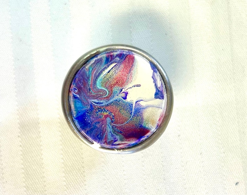May include: A round, silver-coloured knob with a colourful abstract design. The design features swirls of blue, purple, pink, and white paint with a hint of gold glitter.