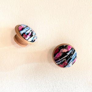 K&ouml;nnte beinhalten: Zwei Holzkn&ouml;pfe mit einem abstrakten Design in Pink, Blau und Schwarz. Die Kn&ouml;pfe sind rund und haben einen erh&ouml;hten Rand.