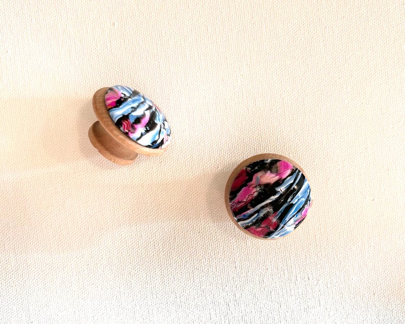 K&ouml;nnte beinhalten: Zwei Holzkn&ouml;pfe mit einem abstrakten Design in Pink, Blau und Schwarz. Die Kn&ouml;pfe sind rund und haben einen erh&ouml;hten Rand.