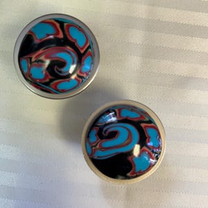 May include: Two round, silver-colored knobs with a swirling blue, black, and red design.