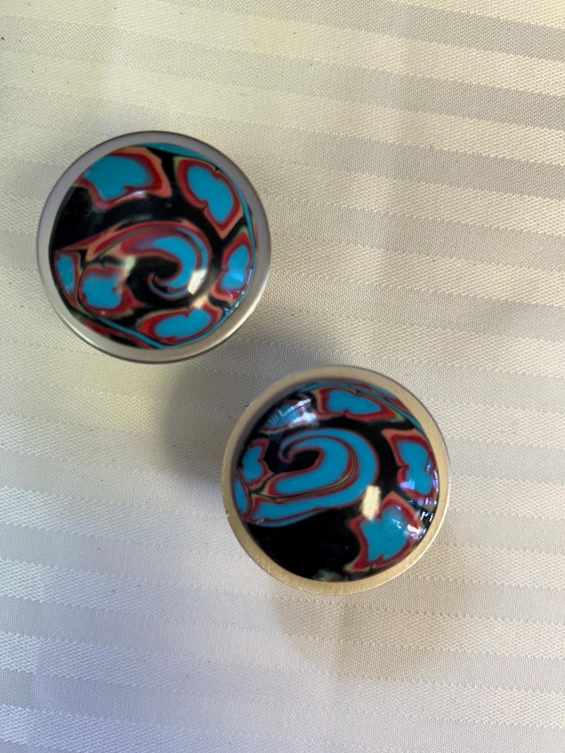 May include: Two round, silver-colored knobs with a swirling blue, black, and red design.