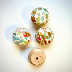 May include: Four round wooden knobs with a white background and colourful abstract patterns. One knob is shown from the bottom, revealing a hole for a screw.