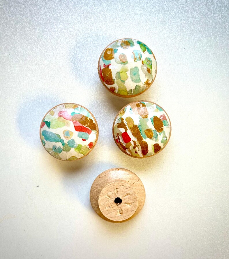 May include: Four round wooden knobs with a white background and colourful abstract patterns. One knob is shown from the bottom, revealing a hole for a screw.