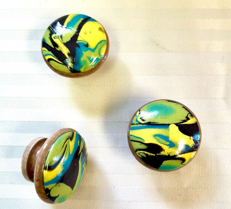 May include: Three decorative cabinet knobs with a colorful, swirled design. The knobs feature a mix of yellow, blue, and black patterns set within a wooden frame. The knobs are round and have a glossy finish.