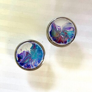 May include: Two round, silver-coloured, decorative knobs with a clear glass top. The glass is filled with a swirling, abstract design of purple, blue, and white paint.