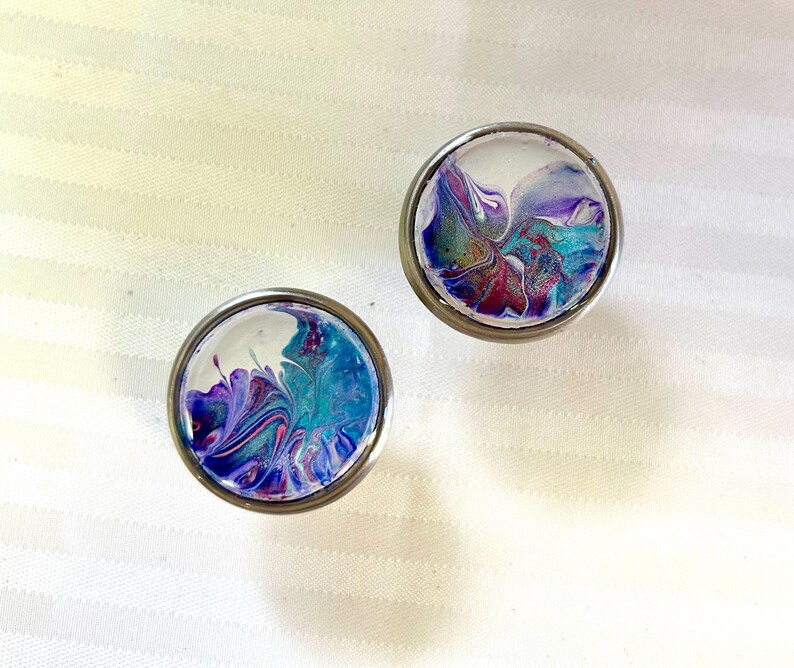May include: Two round, silver-coloured, decorative knobs with a clear glass top. The glass is filled with a swirling, abstract design of purple, blue, and white paint.