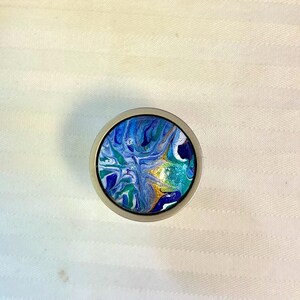 May include: A round, silver-toned bottle stopper with a colorful abstract design. The design features swirls of blue, green, and white paint with gold accents. The stopper is set against a white background.