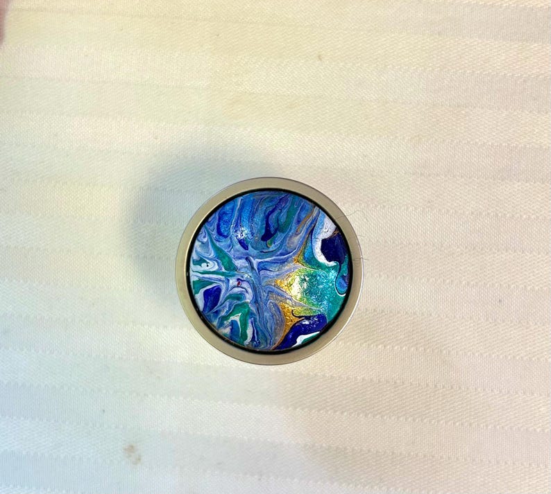 May include: A round, silver-toned bottle stopper with a colorful abstract design. The design features swirls of blue, green, and white paint with gold accents. The stopper is set against a white background.