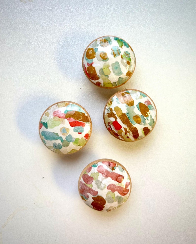 May include: Four round wooden knobs with a white background and abstract patterns in shades of blue, green, red, and gold.