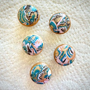 以下が含まれることがあります： カラフルな抽象デザインの丸い木製キャビネットノブ5個。各ノブは、明るい木材ベースに、ティール、ゴールド、ブラック、ピンクのスワールパターンが特徴です。ノブは白いテクスチャのある表面に配置されています。
