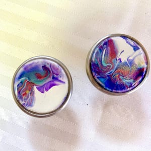 May include: Two round, silver-coloured drawer knobs with a white background and a swirling pattern of purple, blue, and red paint.