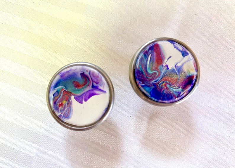 May include: Two round, silver-coloured drawer knobs with a white background and a swirling pattern of purple, blue, and red paint.