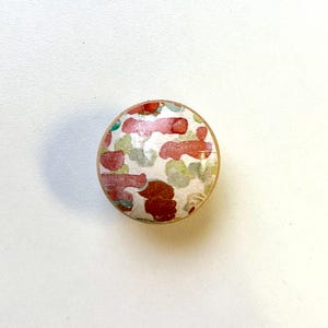 May include: A round, wooden knob with a colourful abstract pattern. The pattern features red, green, and white shapes on a white background.