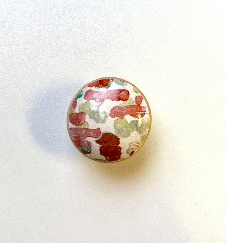 May include: A round, wooden knob with a colourful abstract pattern. The pattern features red, green, and white shapes on a white background.