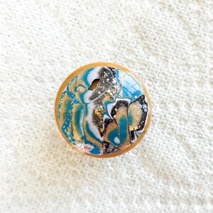 May include: A round wooden knob with a resin top featuring a swirling design in teal, gold, white, and black. The knob has a natural wood edge and is likely for furniture or decor.