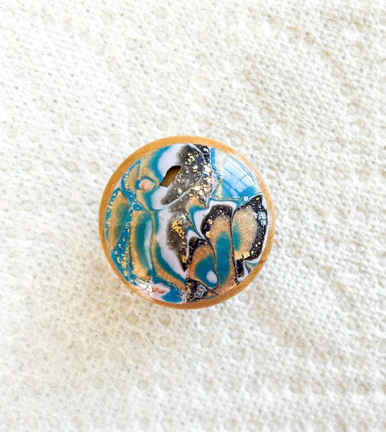 May include: A round wooden knob with a resin top featuring a swirling design in teal, gold, white, and black. The knob has a natural wood edge and is likely for furniture or decor.