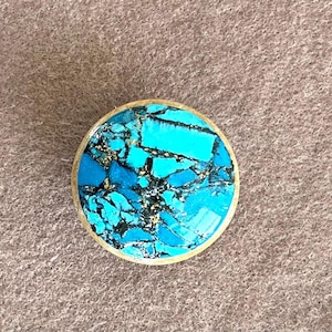 Könnte beinhalten: Ein runder, türkisfarbener und schwarzer Cabochon, der in eine Messingfassung gefasst ist. Der Cabochon hat ein marmoriertes Muster aus Türkis und Schwarz.