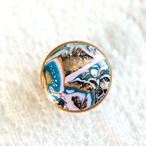 May include: A round, decorative bottle stopper with a wooden base. The top features a swirling design of teal, white, black, and gold, with a light pink accent. The stopper is designed for wine bottles and adds a unique touch.