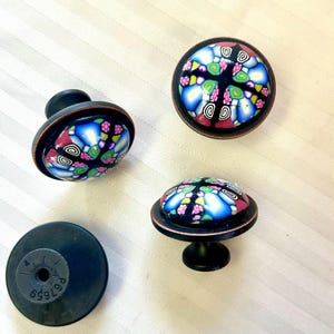 May include: Four decorative cabinet knobs with a colourful, mosaic-like design. The knobs feature a black base and a copper-coloured rim, with a domed, patterned top in shades of blue, pink, green, and black. One knob is shown from the back.