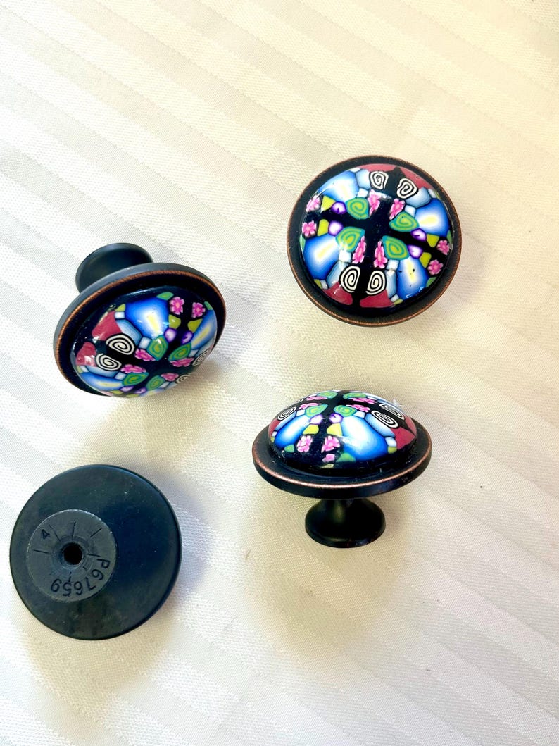 May include: Four decorative cabinet knobs with a colourful, mosaic-like design. The knobs feature a black base and a copper-coloured rim, with a domed, patterned top in shades of blue, pink, green, and black. One knob is shown from the back.
