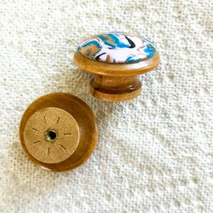 May include: Two wooden cabinet knobs. One knob is upright, showing a colorful, marbled design in blue, white, and gold on top. The other knob is lying on its back, revealing the screw attachment. The knobs are made of light brown wood.