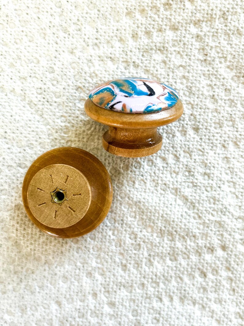 May include: Two wooden cabinet knobs. One knob is upright, showing a colorful, marbled design in blue, white, and gold on top. The other knob is lying on its back, revealing the screw attachment. The knobs are made of light brown wood.