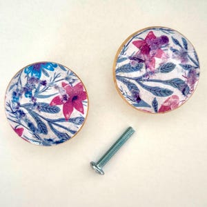 K&ouml;nnte beinhalten: Zwei runde Schrankkn&ouml;pfe mit Blumendekor und einer Schraube. Die Kn&ouml;pfe haben einen wei&szlig;en Hintergrund mit rosa, lila und blauen Blumen und gr&uuml;nen Bl&auml;ttern. Die R&auml;nder der Kn&ouml;pfe sind goldfarben.