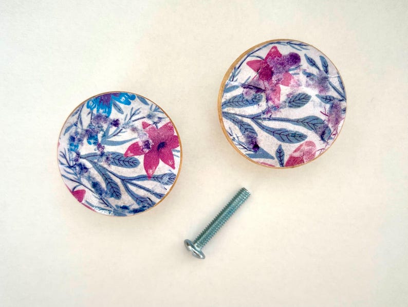 K&ouml;nnte beinhalten: Zwei runde Schrankkn&ouml;pfe mit Blumendekor und einer Schraube. Die Kn&ouml;pfe haben einen wei&szlig;en Hintergrund mit rosa, lila und blauen Blumen und gr&uuml;nen Bl&auml;ttern. Die R&auml;nder der Kn&ouml;pfe sind goldfarben.