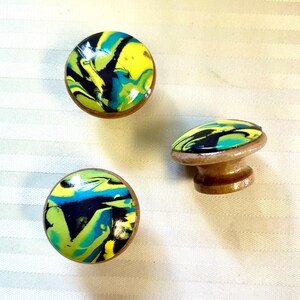 May include: Three decorative cabinet knobs with a colorful, swirled design. The knobs feature a mix of yellow, blue, green, and black patterns on a wooden base. One knob is a standard round shape, and the other is a mushroom shape.