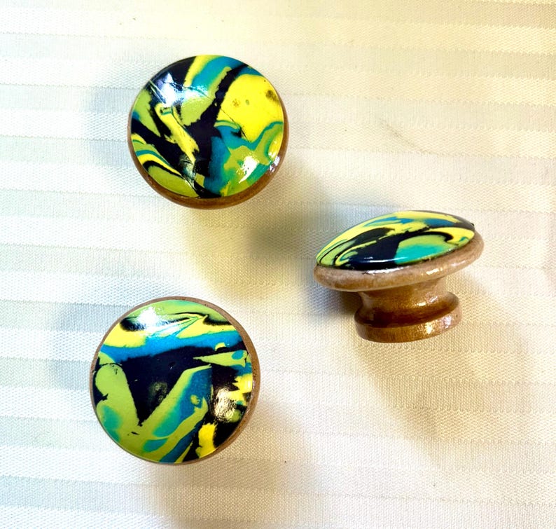 May include: Three decorative cabinet knobs with a colorful, swirled design. The knobs feature a mix of yellow, blue, green, and black patterns on a wooden base. One knob is a standard round shape, and the other is a mushroom shape.