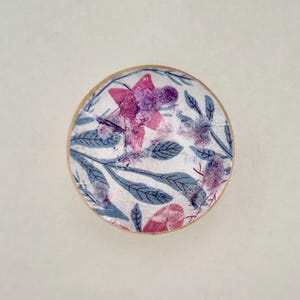 K&ouml;nnte beinhalten: Ein rundes, dekoratives Objekt mit floralem Design. Das Design zeigt rosa und lila Blumen mit blauen Bl&auml;ttern auf wei&szlig;em Hintergrund. Der Rand des Objekts hat einen hellbraunen Holzrahmen.