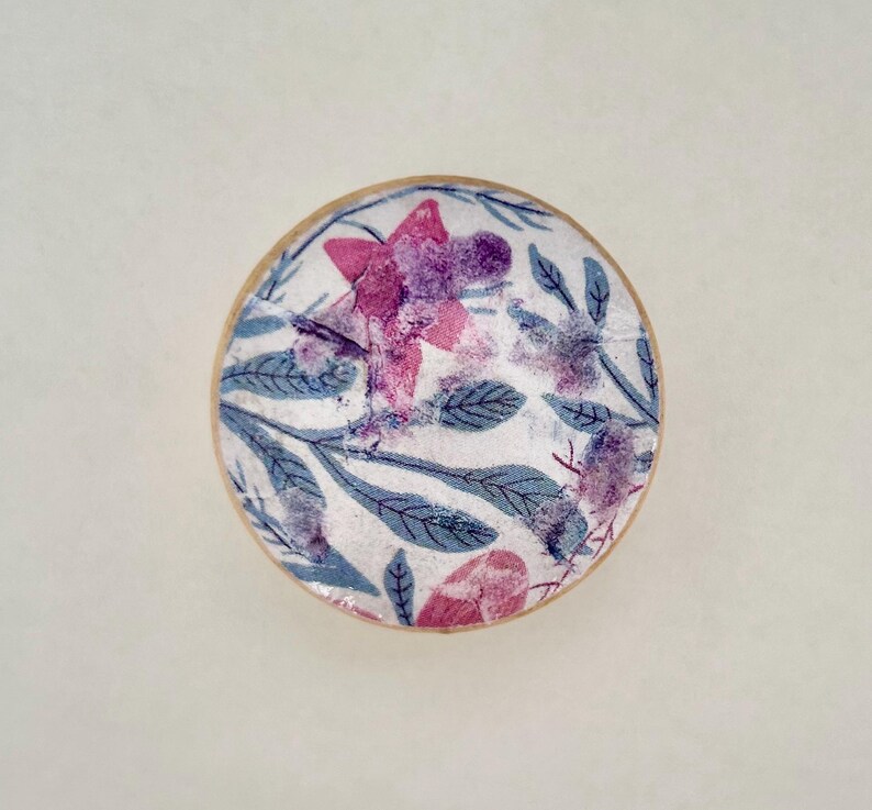 K&ouml;nnte beinhalten: Ein rundes, dekoratives Objekt mit floralem Design. Das Design zeigt rosa und lila Blumen mit blauen Bl&auml;ttern auf wei&szlig;em Hintergrund. Der Rand des Objekts hat einen hellbraunen Holzrahmen.