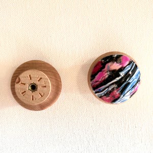 K&ouml;nnte beinhalten: Zwei Holzkn&ouml;pfe mit unterschiedlichen Designs. Ein Knopf hat ein Zifferblattmuster mit r&ouml;mischen Ziffern. Der andere Knopf hat ein farbenfrohes abstraktes Muster mit rosa, blauen und schwarzen Wirbeln.