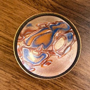 Può includere: Un pomello rotondo in ceramica con un bordo in metallo marrone. Il pomello presenta un motivo a spirale di blu, arancione e bianco.