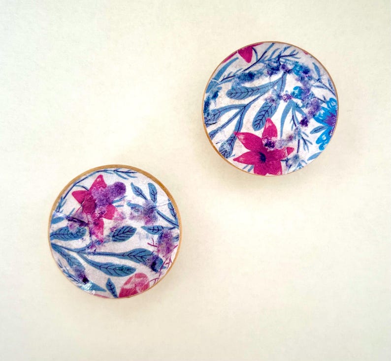 K&ouml;nnte beinhalten: Zwei runde Untersetzer mit floralen Mustern. Die Untersetzer haben einen wei&szlig;en Hintergrund mit rosa, lila und blauen Blumen und Bl&auml;ttern. Die R&auml;nder sind mit hellbraunem Holz eingefasst. Ideal, um jedem Zuhause einen Farbtupfer zu verleihen.