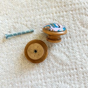 May include: A wooden cabinet knob with a colorful, abstract design on top, a matching wooden base, and a silver screw. The knob has a light pink background with blue, white, and black accents. The base has a starburst pattern.