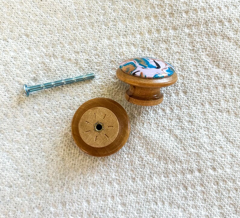 May include: A wooden cabinet knob with a colorful, abstract design on top, a matching wooden base, and a silver screw. The knob has a light pink background with blue, white, and black accents. The base has a starburst pattern.
