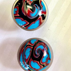 May include: Two round, silver-colored, decorative knobs with a swirling pattern of black, blue, red, and white paint.