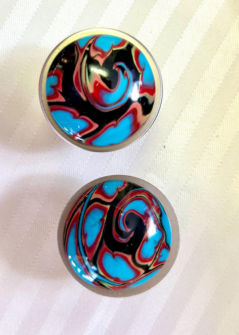 May include: Two round, silver-colored, decorative knobs with a swirling pattern of black, blue, red, and white paint.