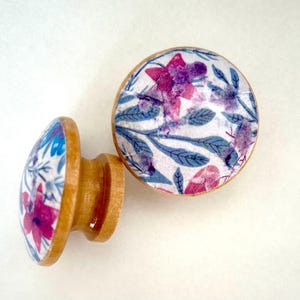 K&ouml;nnte beinhalten: Zwei Holzschrankkn&ouml;pfe mit floralem Design. Die Kn&ouml;pfe haben einen hellbraunen Holzsockel und eine runde Oberseite mit wei&szlig;em Hintergrund und einem Muster aus rosa und lila Blumen und blauen Bl&auml;ttern.