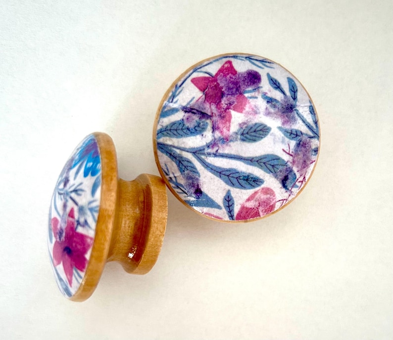 K&ouml;nnte beinhalten: Zwei Holzschrankkn&ouml;pfe mit floralem Design. Die Kn&ouml;pfe haben einen hellbraunen Holzsockel und eine runde Oberseite mit wei&szlig;em Hintergrund und einem Muster aus rosa und lila Blumen und blauen Bl&auml;ttern.